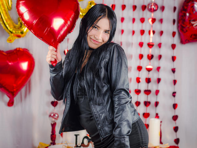 TiffanyEve mit langen, schwarzen Haaren, lächelnd, hält einen glänzenden, roten Herzballon, trägt eine schwarze Lederjacke und ein schwarzes Oberteil und zeigt einen süßen, sexy und kokett wirkenden Look..