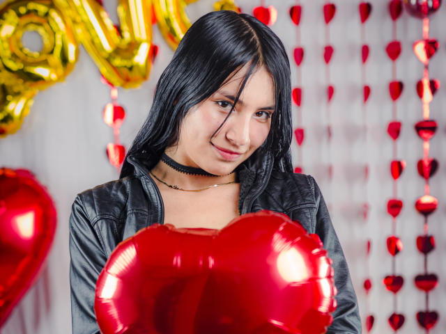 TiffanyEve mit schwarzen Haaren, die eine Lederjacke trägt, rote Herzballons hält und einen schwarzen Choker um den Hals hat, hat ein süßes Lächeln und posiert in einer festlichen, glitzernden Umgebung..
