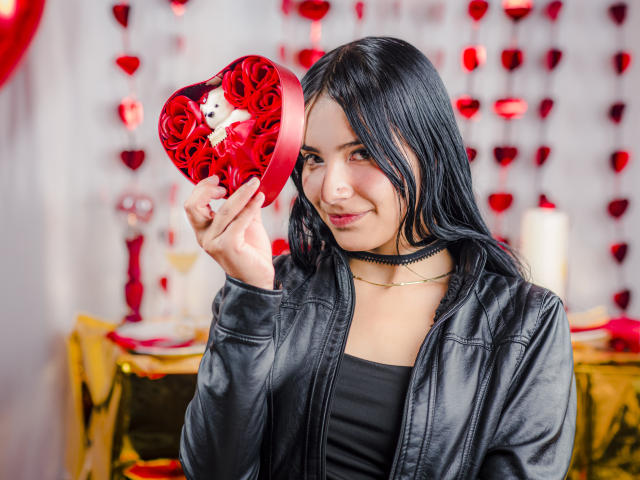 TiffanyEve, con cabello negro, sosteniendo una caja roja en forma de corazón llena de rosas, vistiendo una chaqueta de cuero negra y un collar, sonriendo y mostrando un pequeño gato de peluche blanco..