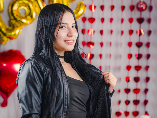 TiffanyEve met lang zwart haar, een zwarte choker, een zwarte leren jas en een zwarte top, die haar haar vasthoudt, lachend, met rode harten en gouden ballonnen op de achtergrond..