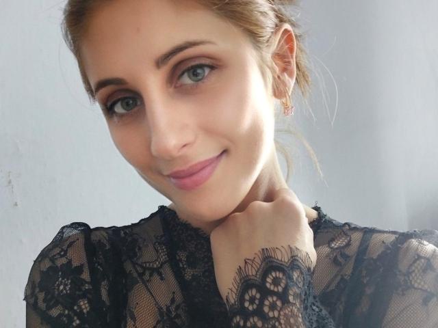 MarinaSexyAndHot, com pele clara, olhos azuis e lábios rosados, vestindo renda preta, com a mão tocando o pescoço, sorrindo gentilmente. Seu cabelo castanho está preso para trás..