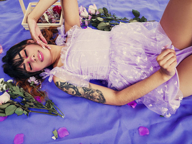 JessicaSoler lies on purple cloth, wearing a sheer lavender dress, showing a tattooed arm, with roses and petals around. Her hand touches her cheek, eyes closed, smiling.