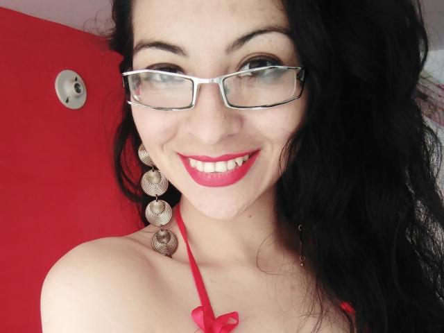 SashaGreish, con i capelli neri, il rossetto rosso e gli occhiali, sorride alla fotocamera, mostrando il seno con una canotta rossa e orecchini a forma di cuore metallici..