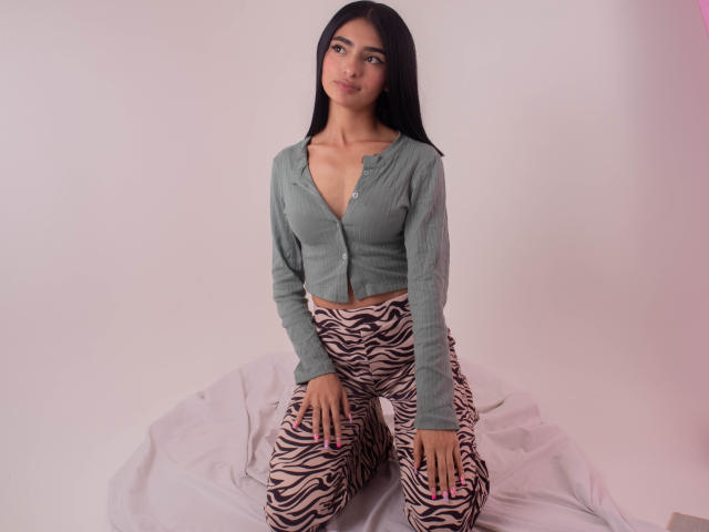 LexiLovie met lang zwart haar, een groene blouse en broek met een zebrapatroon, knielend op een witte doek, lichtelijk opzij kijkend, met een subtiele glimlach..
