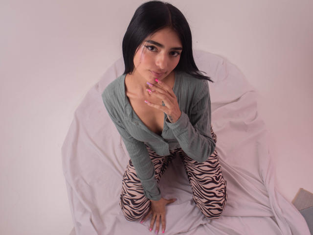 LexiLovie with black hair, green top, zebra pants, and pink nails, kneeling on white sheet, touching her chin, showing cleavage. She has a cute, sexy look.