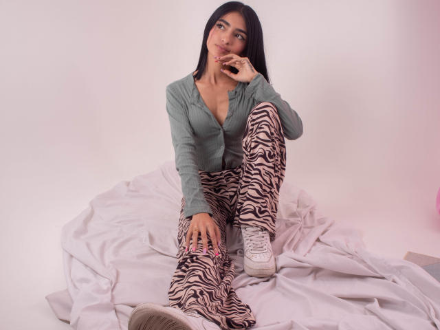 LexiLovie with long black hair, green button-up shirt, zebra pants, and white sneakers, sitting on white fabric, touching her pink lips with a finger.