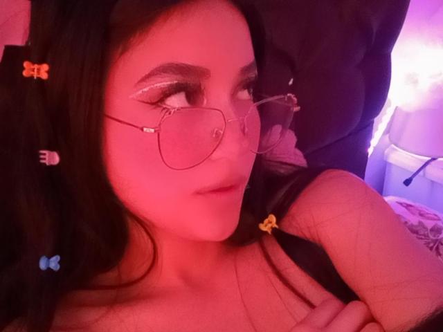 ValeriaClark with pink-tinted skin, black hair, glasses, and a nose ring, looking to the side, wearing a black strap, pink and purple lighting, and small orange hair clips.