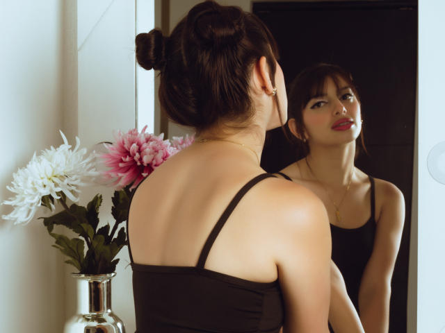 LeticiaFlorenz, avec ses cheveux bruns attachés en chignon, portant un débardeur noir, se reflète dans un miroir. Son rouge à lèvres rouge ressort, et elle regarde son reflet..