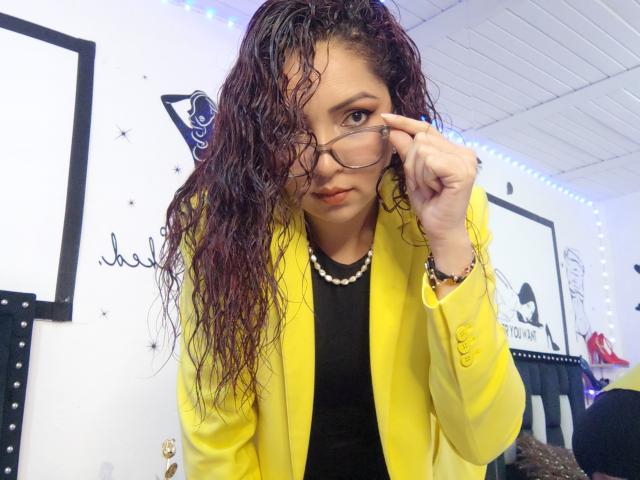 Katrina Bello, com cabelos cacheados e molhados, vestindo um cardigã amarelo, uma blusa preta e um colar de pérolas, ajustando os óculos, com um olhar sedutor, em um quarto bem iluminado..