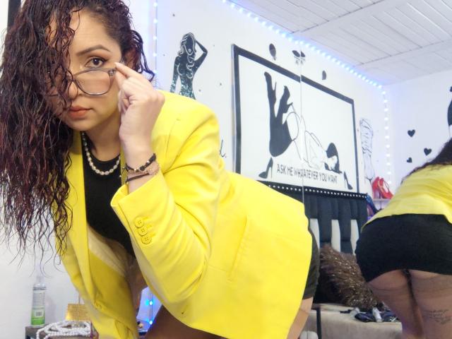 Katrina Bello, avec les cheveux bouclés mouillés, une veste jaune, un t-shirt noir, des lunettes, un collier, penchée en avant, montrant ses fesses dans un miroir, un short noir, une pièce blanche, des lumières LED bleues..