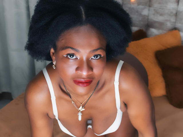 CamilaSandovali with dark skin, afro hair, red lipstick, white bra, necklace with cross, earrings, looking up, on bed, brown and yellow pillows in the background.