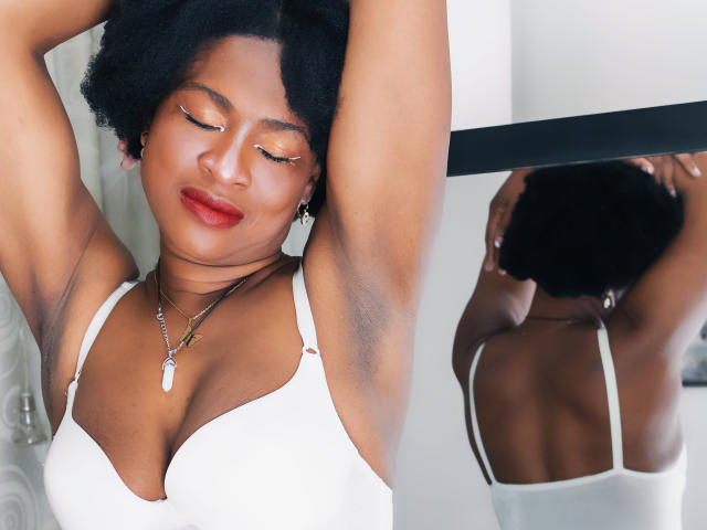 Camila Sandoval, con le braccia alzate e gli occhi chiusi, indossa un reggiseno bianco, mostrando il seno, le ascelle e una collana con un ciondolo bianco, il tutto riflesso in uno specchio..