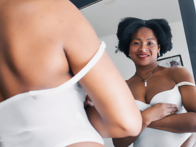 Camila Sandovali, com pele escura e cabelo afro, vestindo uma blusa branca sem alças, olhando no espelho, mostrando seu traseiro e seios pequenos, sorrindo, usando um colar com uma cruz, reflexo no espelho..