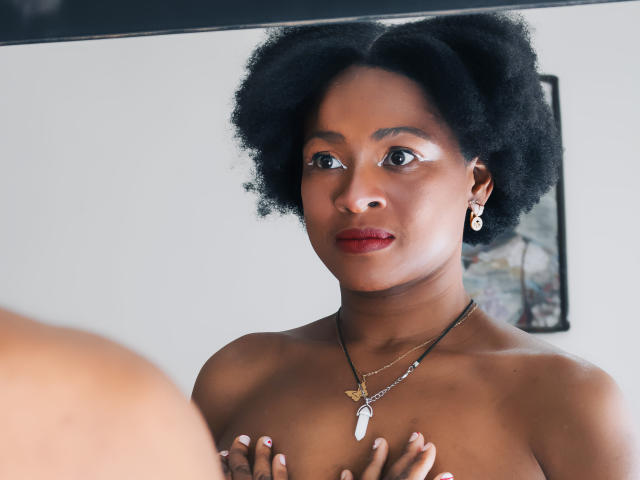 Camila Sandoval, con cabello afro, piel oscura, lápiz labial rojo, usando aretes y collar, cubriendo sus pequeños senos con las manos, mirando en el espejo. Luz natural suave, enfocada en el rostro..