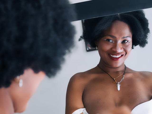 Camila Sandovali, à la peau foncée, avec des cheveux noirs bouclés en afro, souriant en regardant son reflet dans un miroir, portant un collier avec une pierre blanche, nue sur le torse, laissant apparaître ses épaules dénudées..