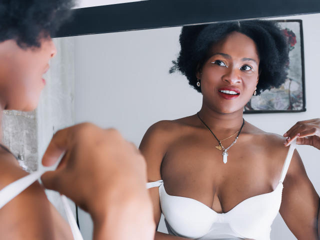 Camila Sandoval, con cabello negro rizado, sonriendo, vestida con un sostén blanco sin tirantes, ajustando una tira, mostrando sus pechos, collar con una piedra blanca, reflejado en un espejo..