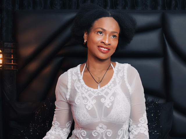 Camila Sandoval, de piel oscura, con cabello afro, vestida con una blusa blanca de encaje, mostrando pequeños senos, sonriendo, con un cabecero de cuero negro, un collar y aretes, en una habitación con luz cálida..