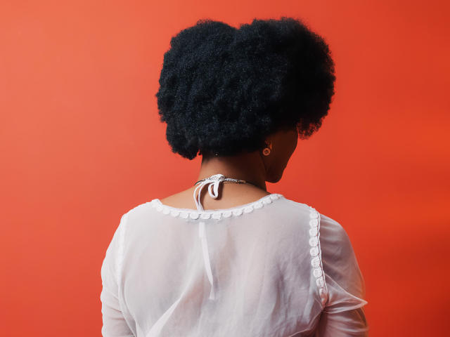 Camila Sandovals schwarze, lockige Haare, sie trägt ein durchsichtiges, weißes Oberteil mit Halstiefe, ein roter Hintergrund, kleine goldene Ohrringe und eine dünne, weiße Kette, sie steht mit dem Rücken zur Kamera..