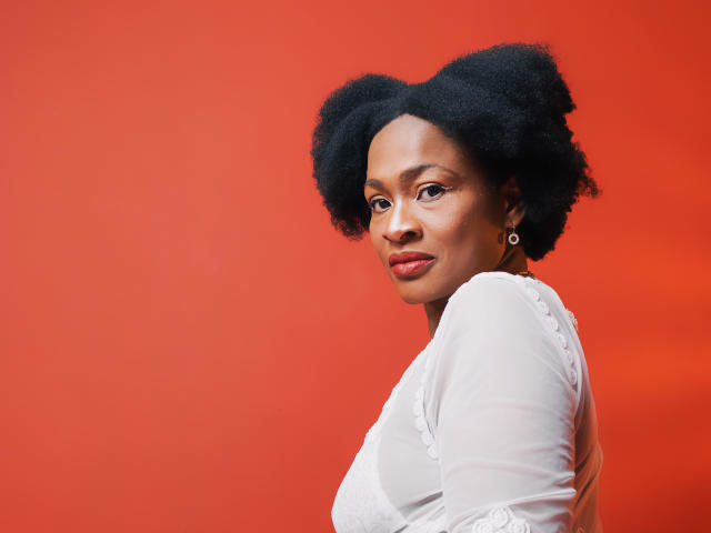 Camila Sandovali, avec une afro noire et volumineuse, portant un haut en dentelle blanche, regardant par-dessus son épaule, fond rouge, sourire discret, petits boucles d'oreilles cerceaux, lumière naturelle douce, confiante, mignonne, attirante..