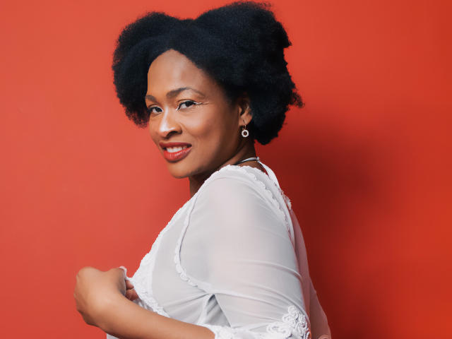 Camila Sandovali con capelli afro neri, top trasparente bianco, rossetto rosso, sorridente alla telecamera, sfondo rosso, piccoli orecchini, che tiene il top, che guarda oltre la spalla, mostrando leggermente il sedere..