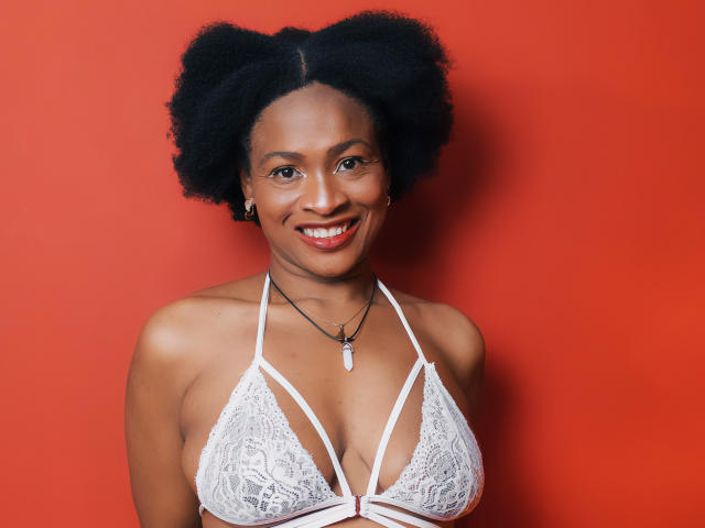 Camila Sandoval, avec ses cheveux noirs et bouclés, souriante, portant un soutien-gorge en dentelle blanche avec des détails apparents, mettant en valeur sa poitrine, sur un fond rouge vif. Elle porte un collier avec un petit pendentif..