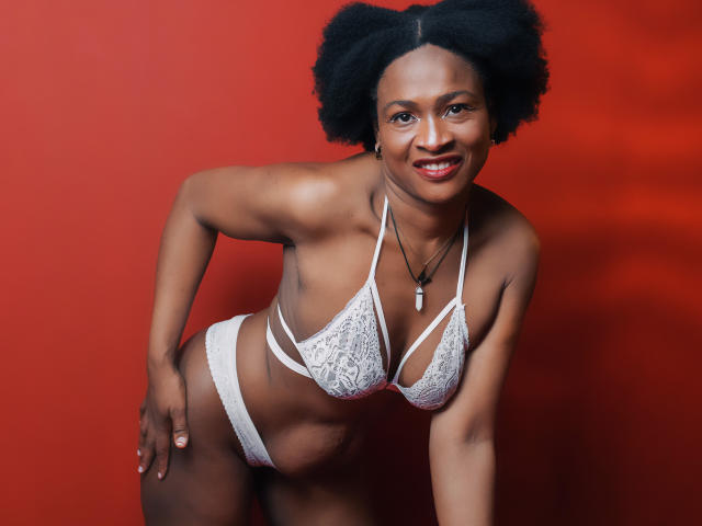 Camila Sandovali, eine schwarze Frau mit Afro, lächelt und trägt einen weißen Spitzen-BH und -Slips. Sie lehnt sich vor und zeigt ihre Brüste vor einem roten Hintergrund. Sie trägt eine Halskette mit einem kleinen Anhänger..