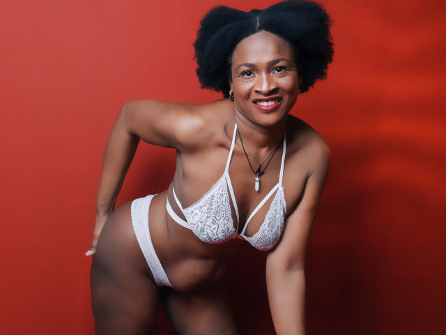 Camila Sandoval, de piel oscura y cabello afro, vestida con un sostén y tanga blancos de encaje, inclinada hacia adelante, sonriendo, mostrando sus pechos y glúteos, sobre un fondo rojo..