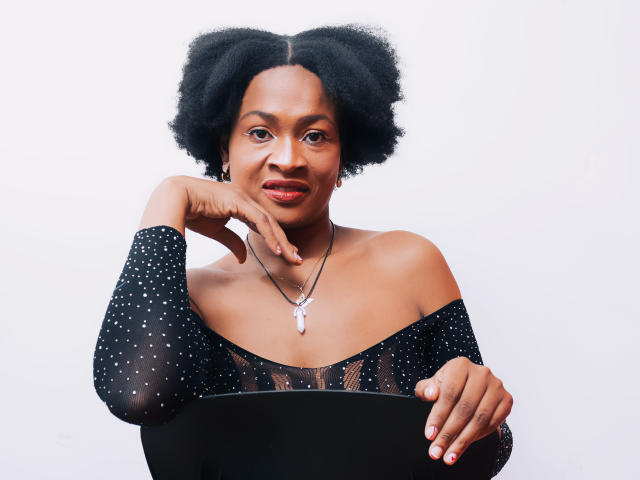 CamilaSandovali with dark skin, black curly hair, wearing a black off-shoulder polka dot top, hand on chin, black necklace with pink stone, smiling subtly.