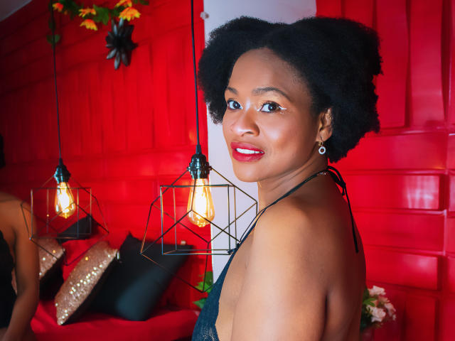 Camila Sandovali, com cabelo afro escuro e encaracolado, batom vermelho, brincos pretos e top de alça, sorrindo para a câmera, com luminárias pretas, uma parede vermelha e almofadas pretas ao fundo..