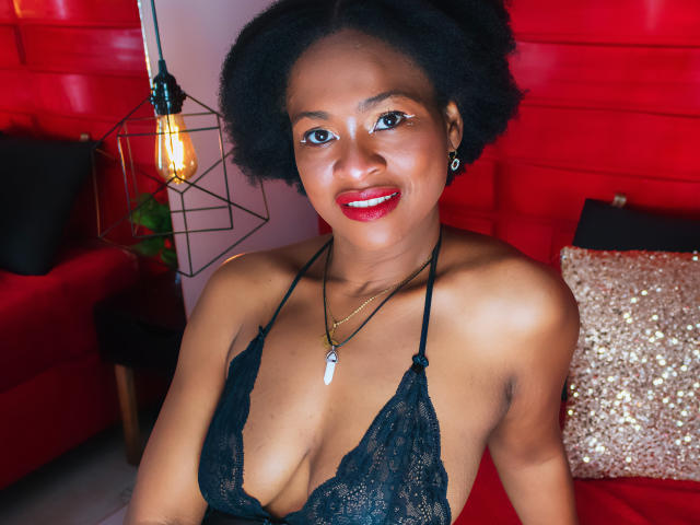 CamilaSandovali with dark skin, short afro, red lipstick, smiling, wearing black lace lingerie, showing her tits, necklace with white pendant, red background, lamp, and red sofa.