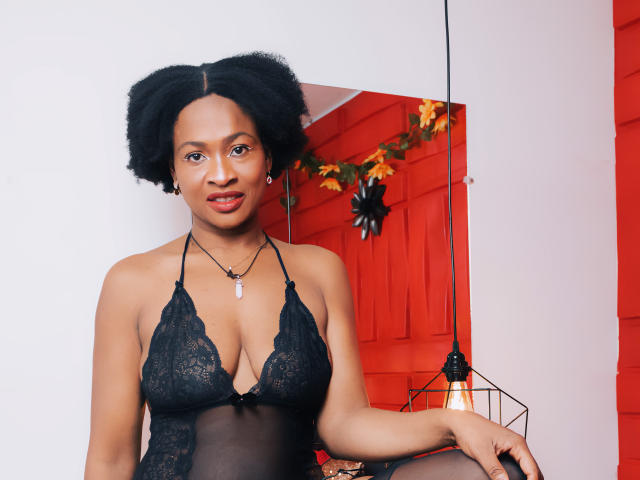 Camila Sandovali mit lockigem, schwarzem Haar, trägt ein schwarzes Spitzen-Dessous-Top, das ihre Brüste zeigt. Sie trägt eine Halskette mit einem weißen Stein und hat einen selbstbewussten Ausdruck..