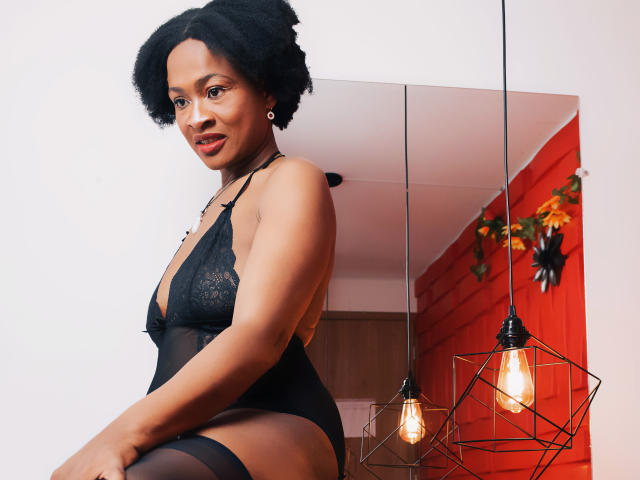 Camila Sandovali in zwart kant lingerie, waarbij haar borsten zichtbaar zijn, met een kort afro kapsel, lachend, staand, met een rode muur, een hangende lamp en een spiegel op de achtergrond. Haar vagina is bedekt met kant..