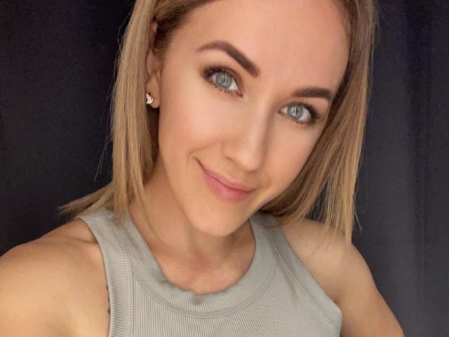 Misstyk has blonde hair, blue eyes, and a cute smile. She's wearing a grey tank top, showing her bare shoulders and small earrings. Her tits are not visible.