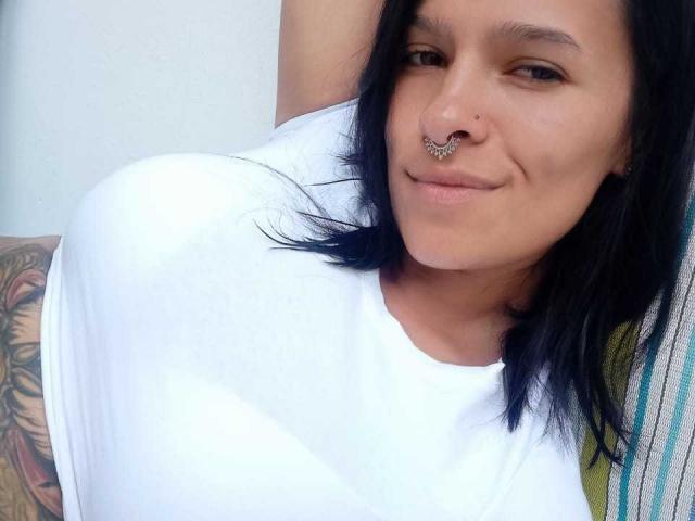 Amelie Blake, con cabello negro, camisa blanca, piercing en la nariz y un brazo tatuado, sonriendo a la cámara, mostrando un busto grande. Piel clara, pose relajada, ligera sonrisa, ambiente casual..