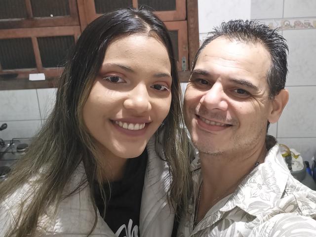 BombomePimenta, com cabelo castanho comprido, nariz pequeno e sorrindo, ao lado de BombomePimenta, com cabelo preto curto, nariz pequeno e sorrindo, em um banheiro, vestindo camisas estampadas, em pé e próximos um do outro..