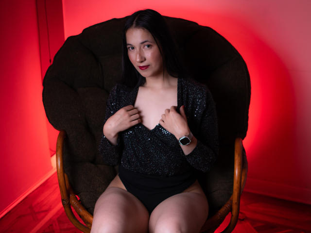 AnneStone with black sparkly top, holding it to show small tits, sitting in chair, black panties, red light, wooden floor, black hair, subtle smile, wearing black watch.
