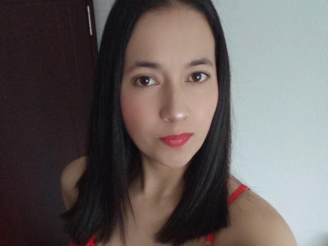 AnneStone has black hair, brown eyes, and red lipstick. She's wearing a red bra, and her medium tits are slightly visible. She's looking directly at the camera.