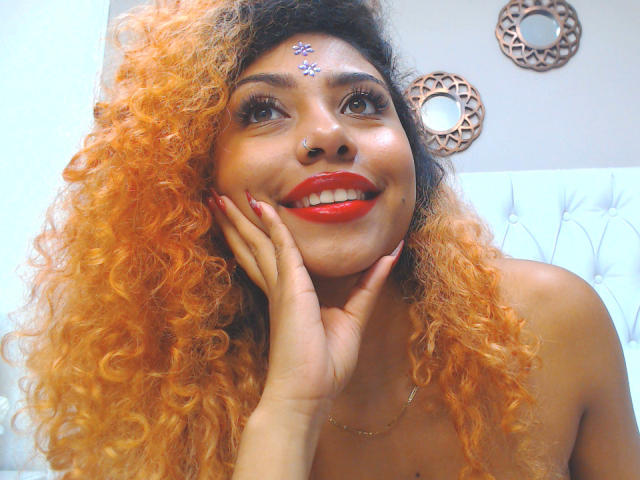 LilithWalker with orange curly hair, red lipstick, nose ring, and decorative forehead star, smiling, hand on cheek, white tufted bed in background.