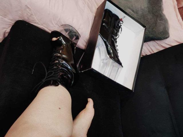 LissaClaire's leg with a black, shiny, lace-up boot, partially on, next to an open box showing the boot. Her pale skin is visible. Boot has a clear heel.