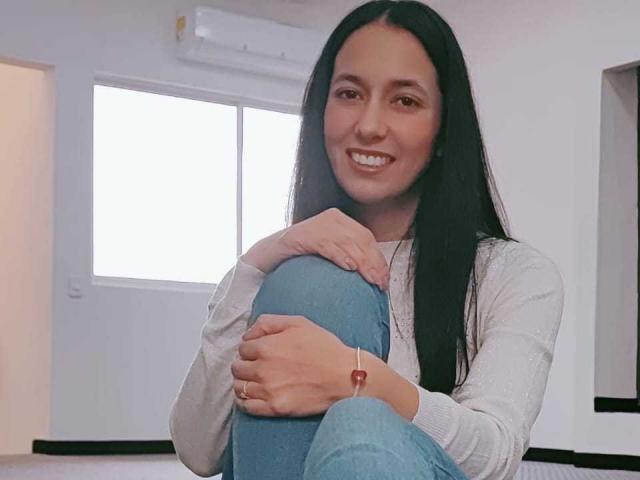 AnneStone with long black hair, smiling, wearing a white sweater and blue jeans, sitting with knees up, hands on knees, red bracelet on right wrist.