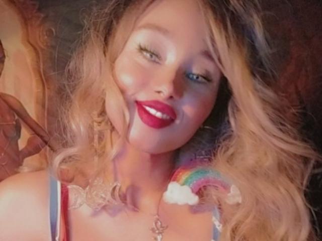 Deusa Nefertari, con i capelli biondi ondulati, rossetto rosso e un arcobaleno e una nuvola sulla spalla, sorride. Indossa una camicetta in pizzo e una collana..