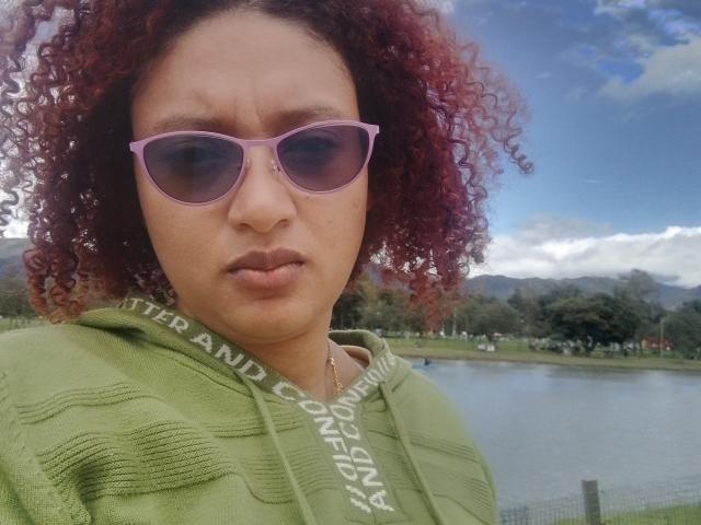VioletaSwitch has red curly hair, pink sunglasses, and a green hoodie, looking slightly annoyed with a pouty face. Her tits are small, and she's outside with a lake.