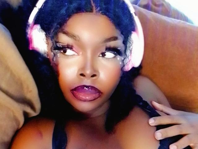 AmandaSykes69, con piel oscura, cabello negro rizado y auriculares rosas, lengua afuera, mirando hacia arriba con pestañas largas, vistiendo una blusa negra, con una mano en el pecho..