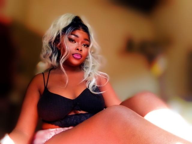 AmandaSykes69 with dark skin, pink lipstick, and platinum blonde curls, wearing a black bra and pink shorts, sitting with legs up, showing her tits and butt.