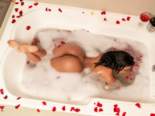 MariamLowel, desnuda, acostada en una bañera blanca llena de agua espumosa y pétalos de rosa roja, con las nalgas levantadas, los pies cruzados, cabello negro ondulado, y una copa de vino tinto a la derecha..