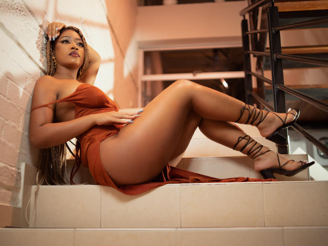 MariamLowel è sdraiata sulle scale, indossa un abito arancione scuro che lascia scoperto il sedere, e porta dei tacchi neri con cinturini. Ha lunghi capelli intrecciati e un'aria sicura e sensuale..