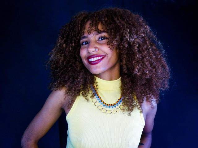 CinthiaWalker with curly brown hair, yellow sleeveless top, blue and orange bead necklace, smiling with red lipstick, nose ring, and dark background. Boobs not visible, focus on face.