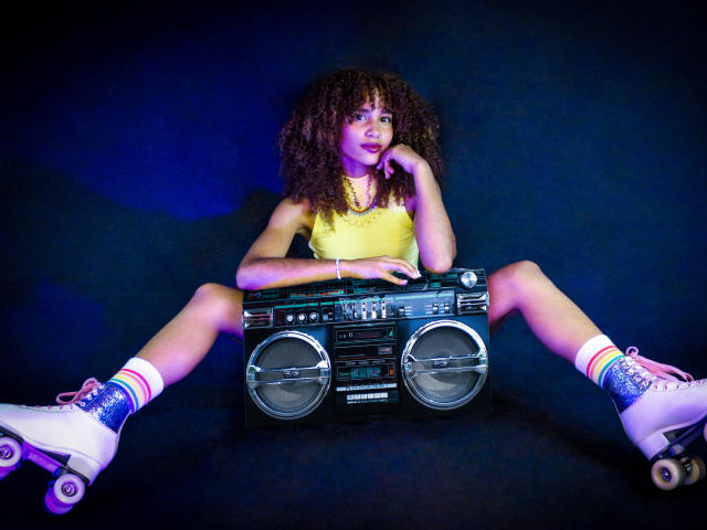 CinthiaWalker with curly brown hair, yellow top, and rainbow socks, sitting with legs spread, holding a boombox, wearing white roller skates, blue and purple lighting.