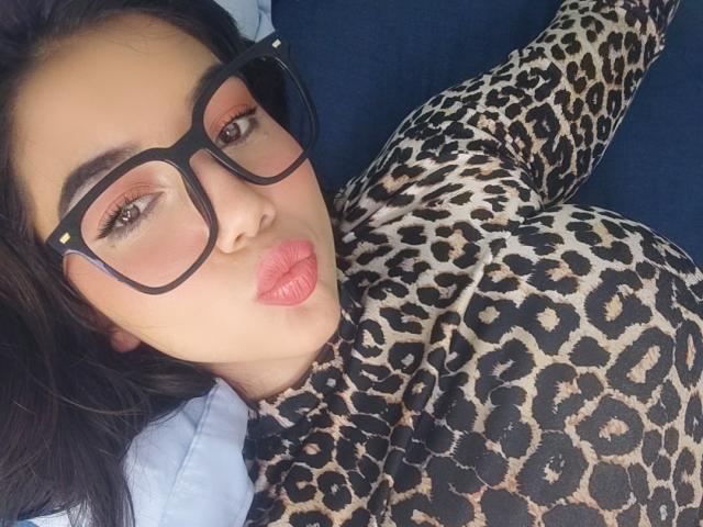 SquirtingCouple with big glasses, pouty pink lips, and leopard print outfit, lying on blue fabric, looking at camera. SquirtingCouple has dark hair and light brown skin.