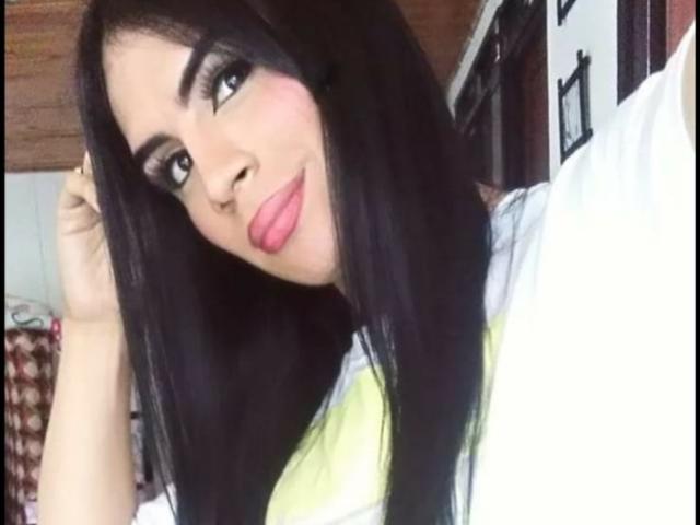 MikeyllaFoxT with long black hair, pink lips, and heavy eye makeup, smiling slightly, hand in hair, wearing a white shirt with yellow stripes.